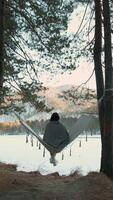 Young woman enjoys peaceful moment, relaxing in hammock by frozen lake, covered with warm blanket, admiring beautiful sunset over snowy landscape and serene winter scenery video