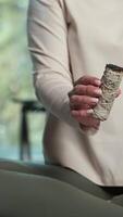 Female practitioner wearing casual attire gently holding white sage smudge stick, slowly rotating to display herbal tool used in spiritual cleansing and energy clearing ritual video