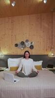 Comfortable room with wooden walls sets calm atmosphere as woman practices mindfulness, meditating in lotus position on bed with laptop open in front of her video