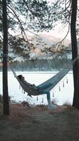 Young woman wearing winter clothes enjoys relaxing in hammock strung between two trees by beautiful frozen lake in snowy winter forest at sunset video