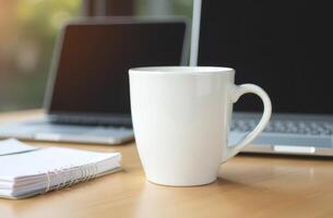 A white coffee mug on a wooden desk with a laptop, notebook, and a blank screen in a bright workspace during the day photo