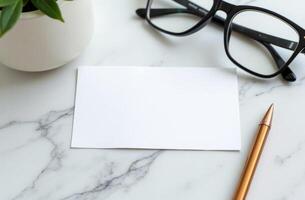 Desk setup featuring a blank card, reading glasses, a gold pen, and a potted plant on a marble surface in soft natural light photo