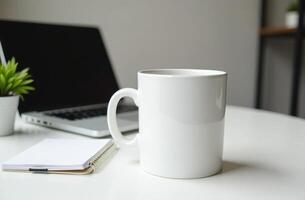A white coffee mug sits on a table beside a laptop and notepad in a minimalistic workspace during daylight photo