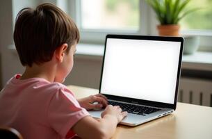 A young child focused on a laptop while sitting at a table in a well-lit room with plants nearby during daylight hours photo