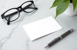 Blank note card beside glasses and pen on a marble desk with a potted plant, ideal for a workspace photo
