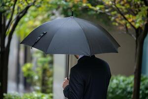 Man holding black umbrella in a rainy urban setting, surrounded by trees and greenery on a cloudy day photo