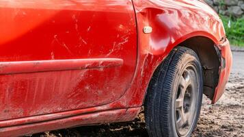 Red car with a damaged side and muddy tires parked on a rough dirt road during late afternoon photo