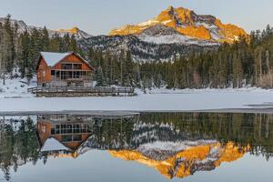 A cabin sits on the shore of a lake with snow covered mountains in the background photo
