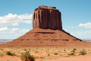 Iconic butte in a desert landscape. A striking, reddish-brown rock formation rises above a vast, arid plain. Sparse vegetation clings to the parched ground. Clear blue sky with a few puffy clouds photo
