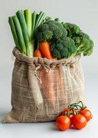 Veggies in a jute bag. A jute bag filled with fresh broccoli, carrots, and green onions sits next to vibrant red tomatoes on a neutral surface. photo