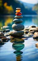 Balanced stones create serene reflection. A stack of smooth stones rests on a riverbank, reflecting in the calm water under a clear sky during daylight. photo