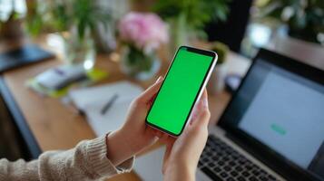Mobile device held in hands with green screen, laptop and workspace in bright indoor setting during daytime photo