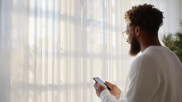 Man in a and sweater uses a smartphone while standing by a white window, surrounded by soft curtains sunlit indoor plants photo