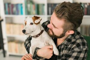 Male owner of jack russell terrier dog, feels responsibility of caring about pet, standing against bookshelves background. People and relationship with animals photo