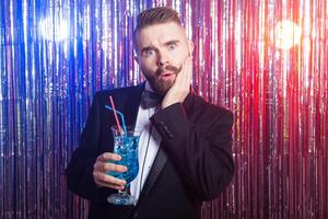 Club party, emotions and holidays concept - Portrait of elegant surprised handsome man in a expensive suit holds blue cocktail on shiny background. photo