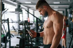 With smartphone, taking ,a break. Strong muscular man is working out in the gym photo