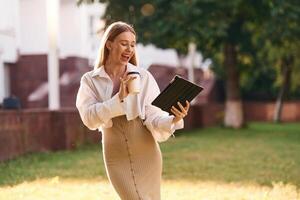 Digital, tablet, and cup of drink in hands. Beautiful female student is outdoors near the university photo