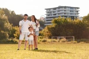 Father, mother and son are standing and holding badminton rackets. On the field photo