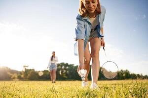 Picking up, the, fallen shuttlecock. Two women are having fun, with badminton rackets, on the field photo