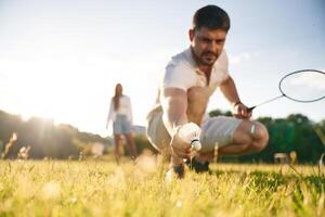 Guy is ,picking up the shuttlecock. Man with woman are playing badminton on the field photo