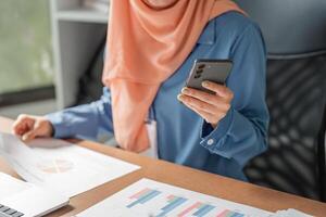 Close-up of Muslim woman in hijab using smartphone while reviewing financial reports at a desk. photo