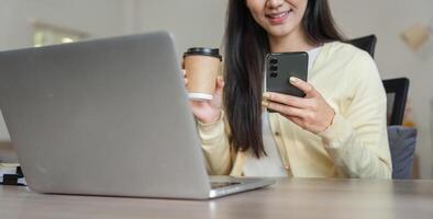 Happy Asian woman enjoying coffee while using smartphone and working on a laptop, reflecting a balanced work-life approach. photo