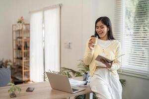 Young woman working from home, enjoying coffee while reading a notebook and smiling. photo
