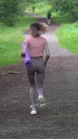 Fit woman running along footpath in park during physical activity, summer workout and physical activity. Rear view of unrecognizable runner wearing sportswear and carries exercise mat. Vertical video