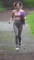 Fit woman running along footpath in park during physical activity, summer workout, physical activity. Front view of slim runner wearing sportswear and carries exercise mat. Full length. Vertical video