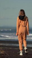 Unrecognizable woman walking along black sandy beach on background of ocean breaking waves at golden hour. Full length back view of model in orange jumpsuit. Vertical format real time cinematic video