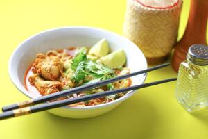 A bowl of spicy noodle soup with chicken, garnished with fresh cilantro lime wedges and chopped vegetables served with black chopsticks. The vibrant colors and presentation are visually appealing. photo