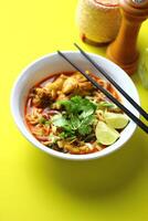 A bowl of spicy noodle soup with chicken, garnished with fresh cilantro, lime wedges, and chopped vegetables served with black chopsticks. The vibrant colors and presentation are visually appealing. photo