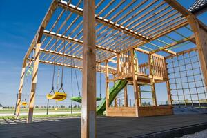 Modern Wooden Playground Structure Featuring Swings, Slide, and Climbing Wall Against a Clear Blue Sky in a Recreational Area photo
