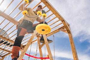 Construction of a Wooden Playground Structure With a Man Installing Yellow Rope Grips in Bright Daylight photo