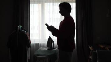 A silhouette of a man standing in front of a window and using a phone video