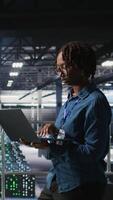 Vertical Happy woman in data center doing maintenance tasks on laptop, ensuring optimal performance. Engineering team reviewing diagnostics and applying patches to server room, improving stability, camera A video