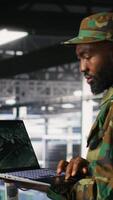 Vertical Military technician in data center monitoring warfare drone telemetry on laptop dashboard. IT specialist in army server room using tactical interface on notebook to support aerial operations, camera B video