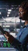 Vertical Data center engineer wearing headset microphone providing remote diagnostics using tablet. Technician monitors system health and guides users through infrastructure troubleshooting steps, camera A video