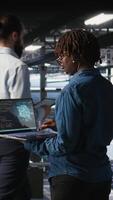 Vertical System administrators in data center monitoring AI LLM visualization. African american server farm coworkers overseeing rigs powering machine learning processes, camera A video
