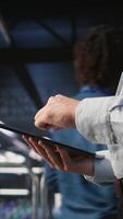Vertical Close up of server room worker checking code on tablet next to colleague reviewing AI LLM visualization. Data center IT expert using device to monitor artificial intelligence systems, camera A video