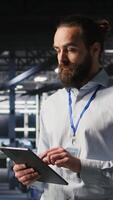 Vertical Cheerful data center admin doing maintenance tasks on tablet, ensuring optimal performance. Upbeat IT professional reviewing diagnostics and applying patches to server hub gear, camera A video