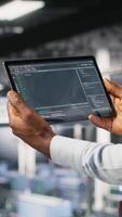 Vertical Data center admin checking code on tablet next to colleague reviewing AI visualizations in blurry background. IT experts use device to analyze neural networks outputs, ensuring stable gear operations video