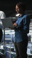Vertical Data center system administrators doing maintenance tasks on laptop, ensuring optimal performance. IT specialists cooperating to review diagnostics and applying patches to server hub gear, camera A video