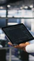 Vertical Close up of data center admin using tablet to reviewing machine learning code dashboards. Server farm IT expert using device to monitor AI infrastructure performance, camera B video