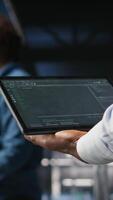 Vertical Close up of data center admin using tablet to reviewing machine learning code dashboards. Server farm IT expert using device to monitor AI infrastructure performance, camera A video