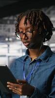 Vertical Server room admin using tablet and headset microphone to provide infrastructure tech support. Data center IT specialist resolves backend issues while coordinating with remote teams, camera B video