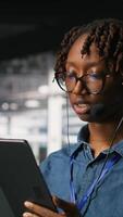 Vertical Data center engineer wearing headset microphone providing remote diagnostics using tablet. Technician monitors system health and guides users through infrastructure troubleshooting steps, camera B video
