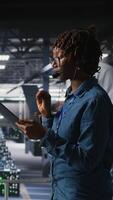 Vertical Server room admin using tablet and headset microphone to provide infrastructure tech support. Data center IT specialist resolves backend issues while coordinating with remote teams, camera A video