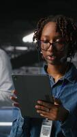 Vertical Data center employee talks in headset microphone while overseeing maintenance on artificial intelligence rigs below. IT professional uses tablet to coordinate machine learning system tasks with team video