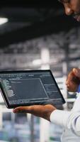 Vertical Close up of programmer swiping on tablet display in data center, doing maintenance tasks, ensuring optimal performance. IT expert checks code on device, applying patches to improve stability, camera B video